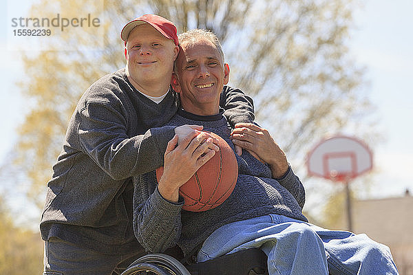 Porträt eines glücklichen Vaters mit Rückenmarksverletzung und seines Sohnes mit Down-Syndrom  der im Park einen Basketball hält