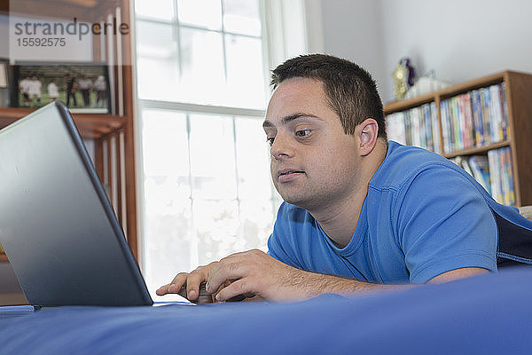 Mann mit Down-Syndrom liegt auf einem Bett und benutzt einen Laptop