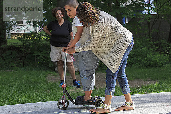 Hispanischer Junge mit Autismus  der mit seiner Mutter und Großmutter im Park auf einem Roller spielt