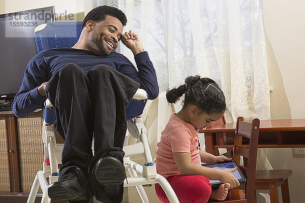 Glücklicher afroamerikanischer Mann mit zerebraler Lähmung und seiner Tochter  die zu Hause ein Tablet benutzt