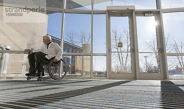 Arzt mit Muskeldystrophie im Rollstuhl am Krankenhauseingang drückt auf ein zugängliches Türschild