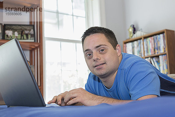 Mann mit Down-Syndrom liegt auf einem Bett und benutzt einen Laptop
