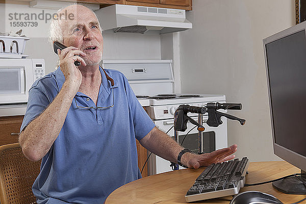 Mann mit Ataxie spricht mit seinem Handy am Computer in der Küche