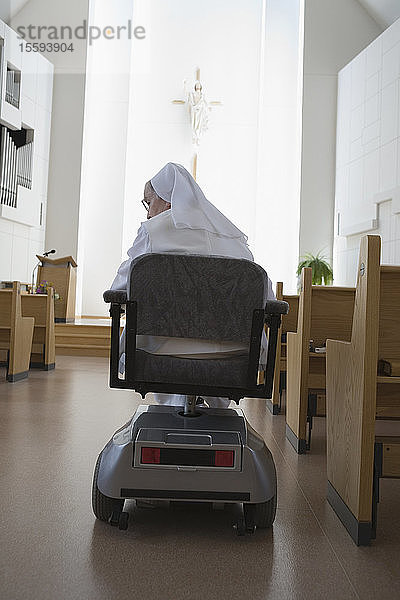 Rückansicht einer in einem motorisierten Rollstuhl sitzenden Nonne in einer Kirche