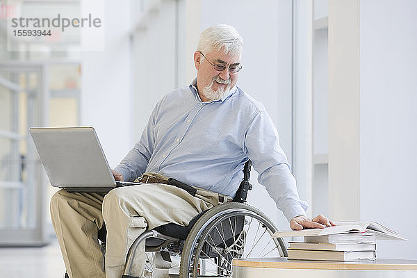 Universitätsprofessor mit Muskeldystrophie liest ein Buch und benutzt einen Laptop