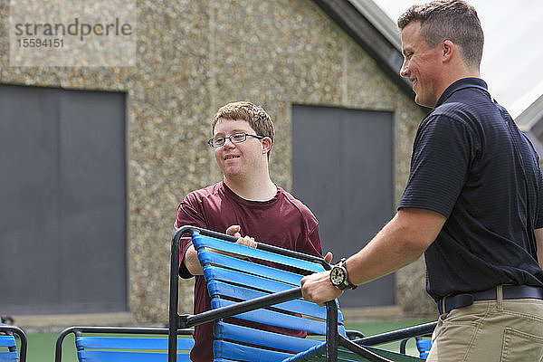 Junger Mann mit Down-Syndrom  der mit einem Betreuer einen Stuhl auf einem College-Ballplatz bewegt