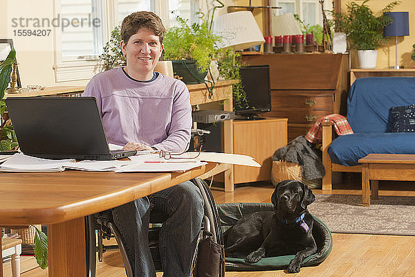 Geschäftsfrau mit Multipler Sklerose im Rollstuhl erledigt zu Hause Papierkram zusammen mit ihrem Diensthund