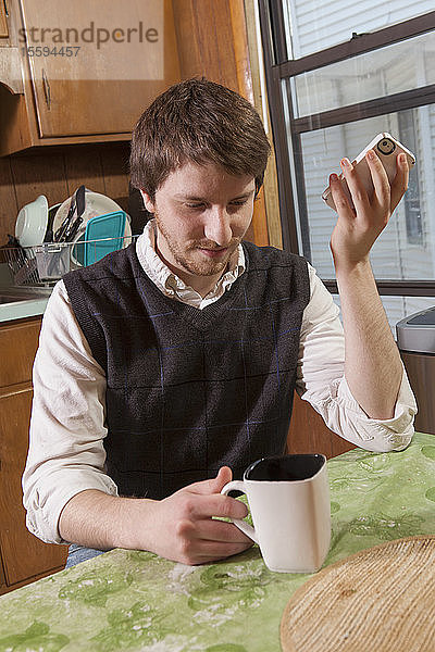 Blinder Mann in seiner Küche  der beim Kaffeetrinken Hilfsmittel auf seinem Mobiltelefon hört