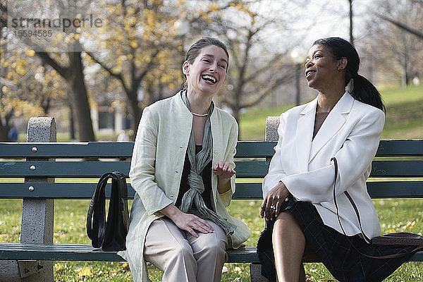 Zwei Frauen sitzen auf einer Bank im Park und lachen.