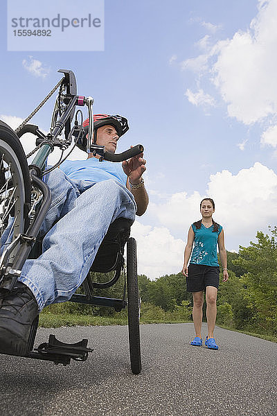 Junger Mann mit einer Rückenmarksverletzung fährt mit einem adaptiven Fahrrad  im Hintergrund steht eine junge Frau