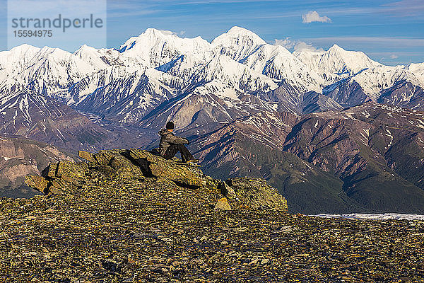 Ein Wanderer  der sich auf einem Felsen ausruht  beobachtet McGinnis Peak  Mount Moffit und Mount Hayes  die sich in der Ferne erheben; Alaska  Vereinigte Staaten von Amerika