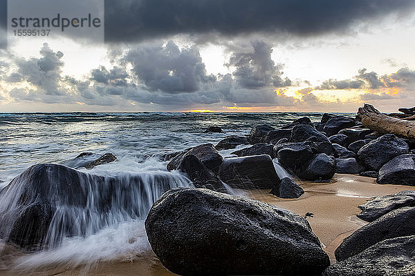 Die Brandung spült über die Felsen am Strand  während die Sonne über dem Ozean mit dunklen Wolken über dem Kopf aufgeht; Kapaa  Kauai  Hawaii  Vereinigte Staaten von Amerika