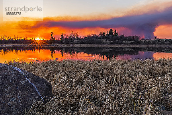 Eine Rauchwolke  die vom Oregon Lakes-Feuer 2019 aufsteigt  spiegelt sich bei Sonnenuntergang in einem See südlich von Delta Junction; Alaska  Vereinigte Staaten von Amerika