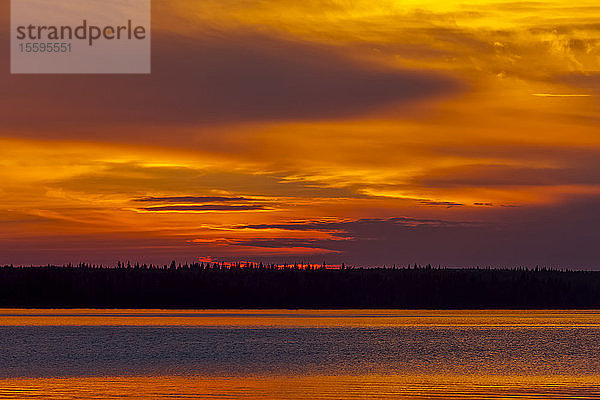 Dramatischer glühender Himmel bei Sonnenuntergang über einem See und einer silhouettierten Uferlinie; Saskatchewan  Kanada