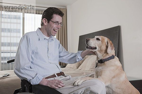 Ein Mann im Rollstuhl mit einer Rückenmarksverletzung spielt mit einem Diensthund im Schlafzimmer