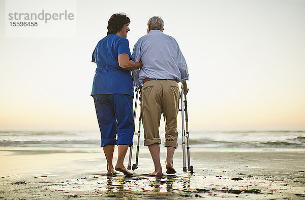 Ein älterer Mann geht mit einer Gehhilfe neben einer Krankenschwester am Strand