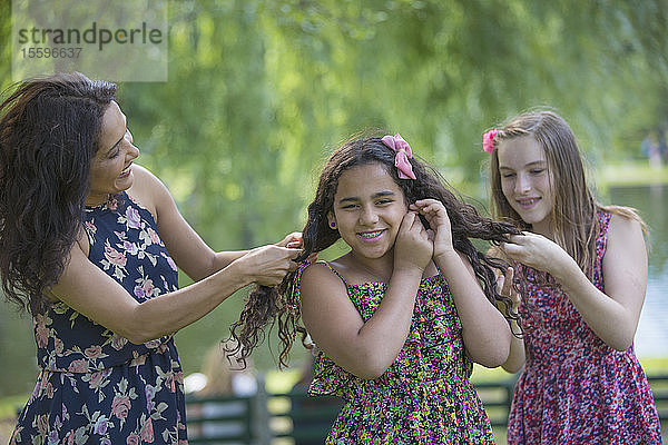 Glückliche hispanische Mutter mit zwei Teenager-Töchtern mit Zahnspange  die sich im Park die Haare machen