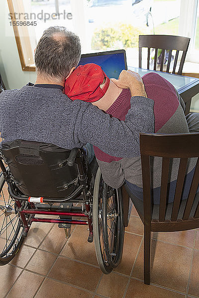 Junger Mann mit Down-Syndrom umarmt seinen Vater im Rollstuhl mit Rückenmarksverletzung zu Hause