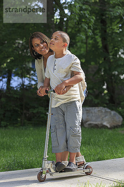 Hispanischer Junge mit Autismus  der mit seiner Mutter im Park auf einem Tretroller spielt