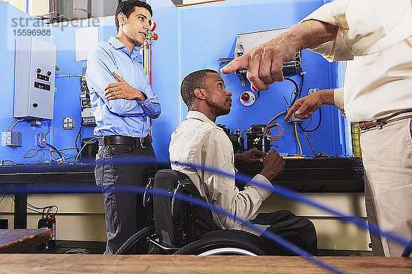 Ausbilder bespricht Kondensatorspule eines Kühlgeräts mit einem Studenten im Rollstuhl