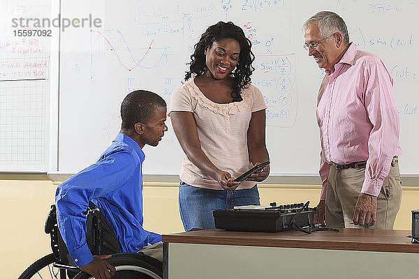 Zwei Studenten der Ingenieurswissenschaften  einer im Rollstuhl  betrachten ein elektronisches Tablet mit einem Professor in einem Klassenzimmer