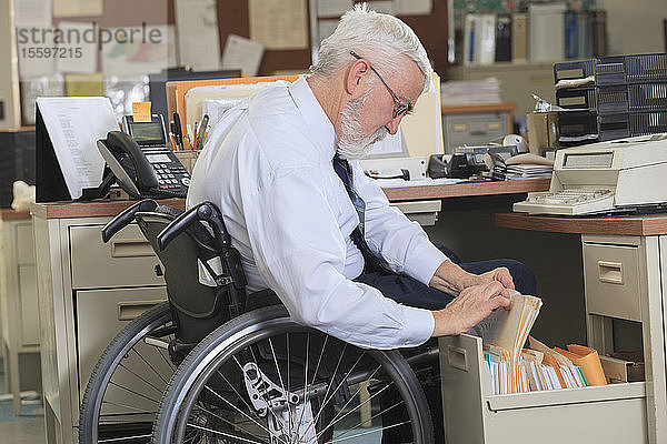 Mann mit Muskeldystrophie im Rollstuhl  der in der Schublade seines Büros Papierkram nachschlägt