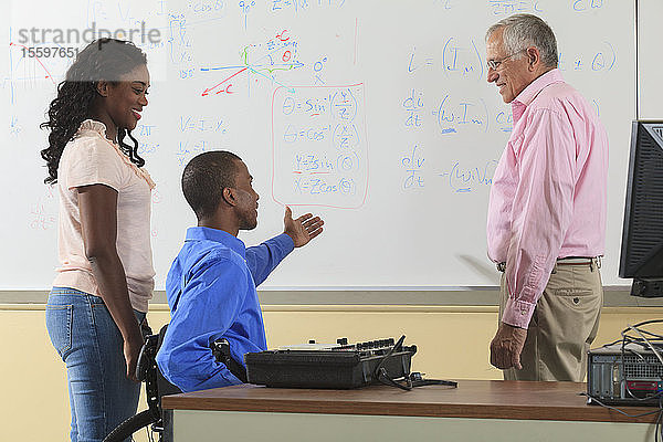 Elektronikprofessor bespricht mit Studenten der Ingenieurwissenschaften  von denen einer im Rollstuhl sitzt  eine Gleichung auf einem Whiteboard