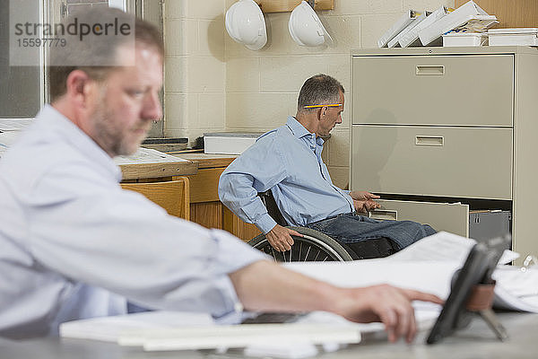Zwei Projektingenieure arbeiten im Büro auf der Baustelle  einer sitzt im Rollstuhl und hat eine Rückenmarksverletzung