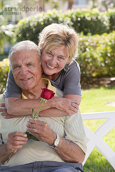 Porträt einer älteren Frau  die einen älteren Mann von hinten mit einer Rose umarmt