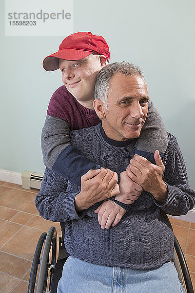 Porträt eines glücklichen jungen Mannes mit Down-Syndrom und seines Vaters im Rollstuhl mit Rückenmarksverletzung zu Hause