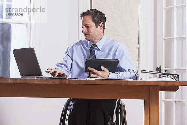 Geschäftsmann mit Querschnittslähmung im Rollstuhl  der ein Tablet und einen Laptop benutzt