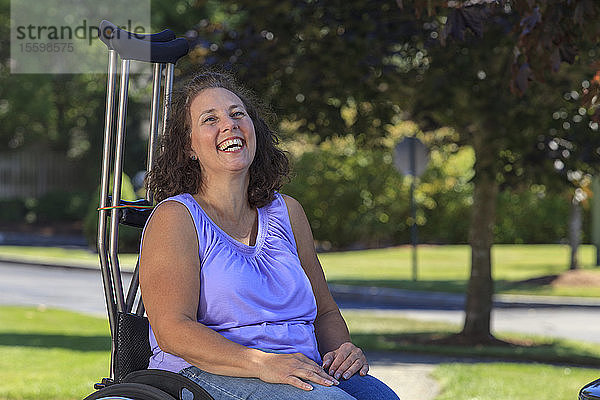 Frau mit Spina Bifida lacht  während sie mit Krücken im Rollstuhl sitzt