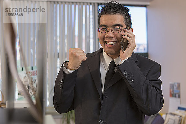 Fröhlicher asiatischer Mann mit Autismus  der mit seinem Handy in einem Büro jubelt