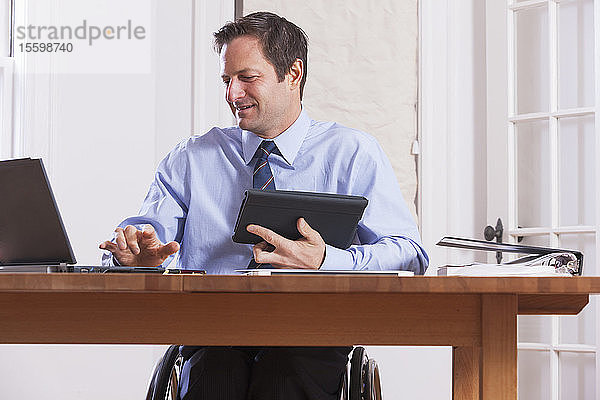 Geschäftsmann mit Querschnittslähmung im Rollstuhl  der ein Tablet und einen Laptop benutzt