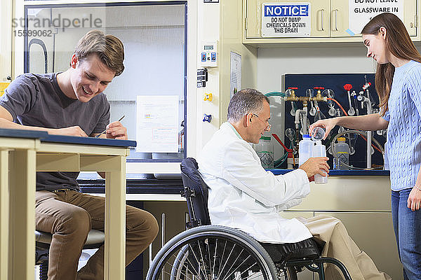 Ein Professor mit einer Rückenmarksverletzung im Rollstuhl arbeitet mit Studenten in einem technischen Labor und füllt einen Behälter mit destilliertem Wasser