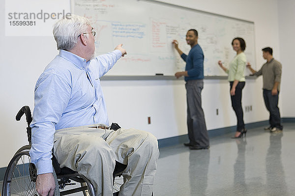 Universitätsprofessor mit Muskeldystrophie im Rollstuhl zeigt auf seine Studenten  die auf eine Tafel schreiben