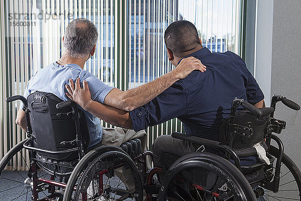 Zwei Männer mit Rückenmarksverletzungen in Rollstühlen  die durch ein Bürofenster hinausschauen und die Arme um sich legen