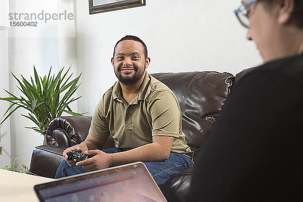 Glücklicher afroamerikanischer Mann mit Down-Syndrom  der einen Game-Controller benutzt  während seine Mutter zu Hause einen Laptop benutzt