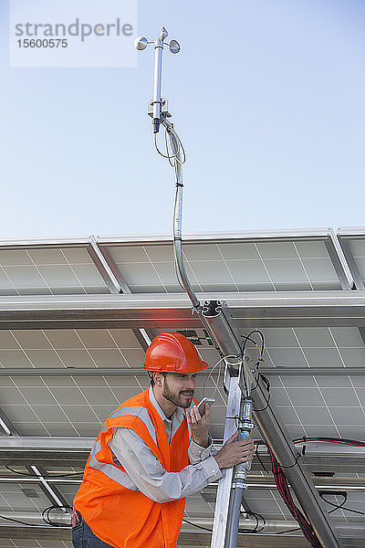 Ein Energietechniker telefoniert mit einem Handy  während er die Leitung der Wetterstation an der Photovoltaikanlage hält.