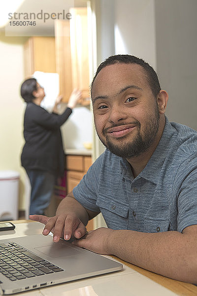 Glücklicher afroamerikanischer Mann mit Down-Syndrom  der zu Hause einen Laptop benutzt  mit seiner Mutter im Hintergrund