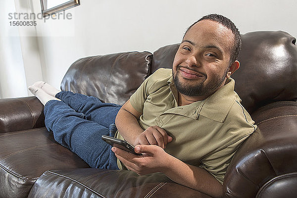 Glücklicher afroamerikanischer Mann mit Down-Syndrom  der zu Hause auf dem Sofa ein Mobiltelefon benutzt