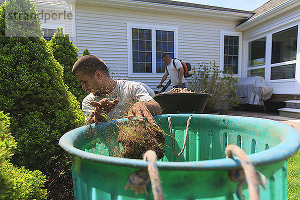 Landschaftsgärtner  die in einem Hausgarten Unkraut in eine Tonne räumen und ein Gebläse zur Reinigung verwenden