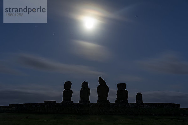 Eine Reihe von Moais hebt sich als Silhouette vom Blau des Nachthimmels ab. Der diffuse Vollmond ist am oberen Rand des Bildes zentriert; Osterinsel  Chile