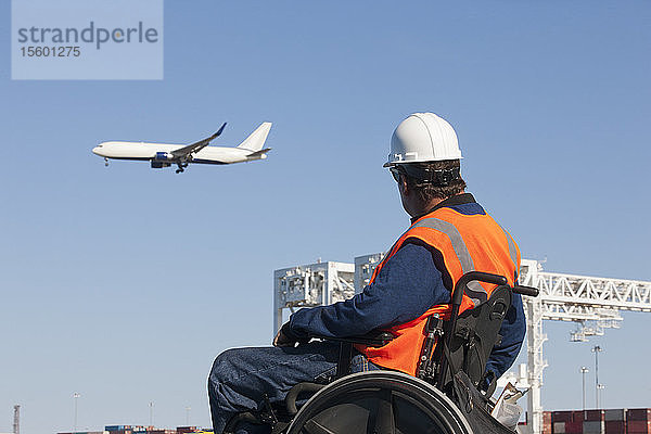 Verkehrsingenieur im Rollstuhl beobachtet ein Flugzeug  das über Schiffscontainer und Kräne im Hafen fliegt