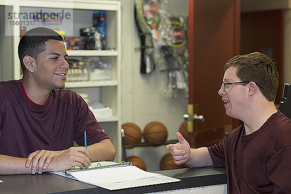 Junger Mann mit Down-Syndrom arbeitet in der Ausgabestelle für Sportgeräte an der Hochschule und spricht mit seinem Vorgesetzten