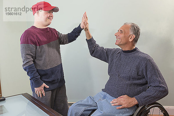 Glücklicher junger Mann mit Down-Syndrom und sein Vater im Rollstuhl mit Rückenmarksverletzung haben Spaß zu Hause