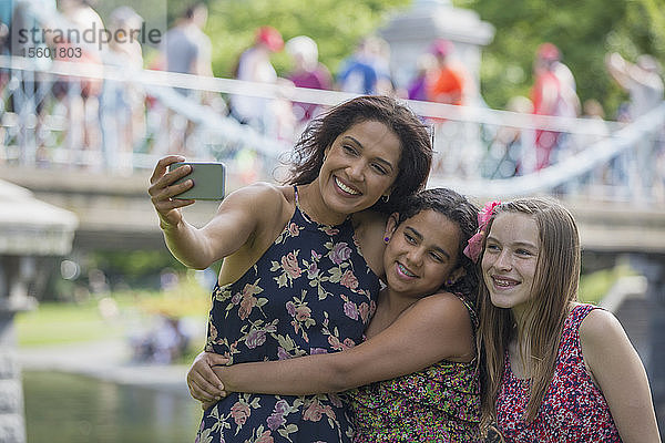 Glückliche hispanische Mutter mit zwei Teenager-Töchtern  die ein Selfie mit ihrem Handy im Park machen