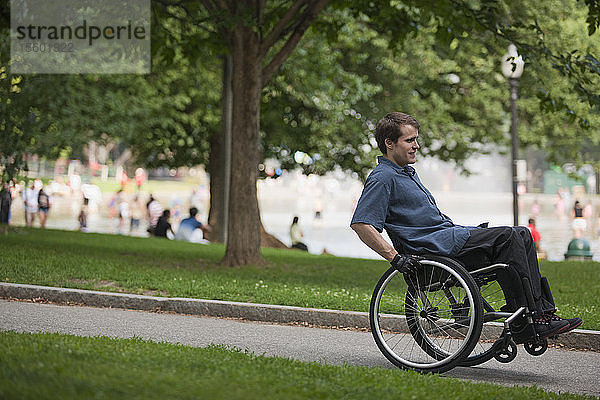 Mann mit Querschnittslähmung im Rollstuhl auf einem Weg in einem öffentlichen Park