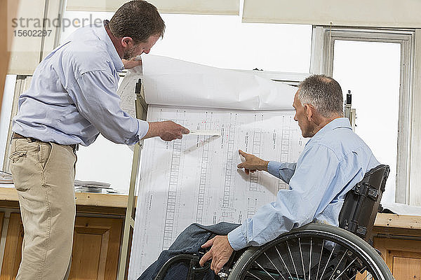 Zwei Projektingenieure benutzen ein Hängeregal für Baustellenpläne  einer sitzt im Rollstuhl und hat eine Wirbelsäulenverletzung