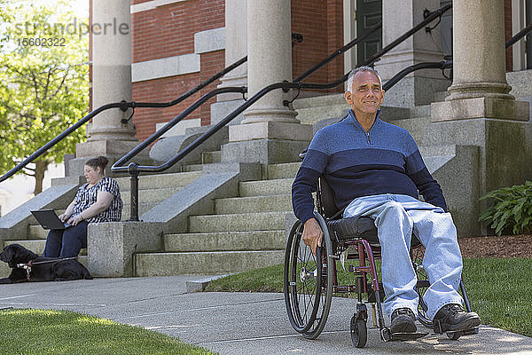 Mann mit Rückenmarksverletzung sitzt im Rollstuhl mit seiner blinden Tochter im Hintergrund